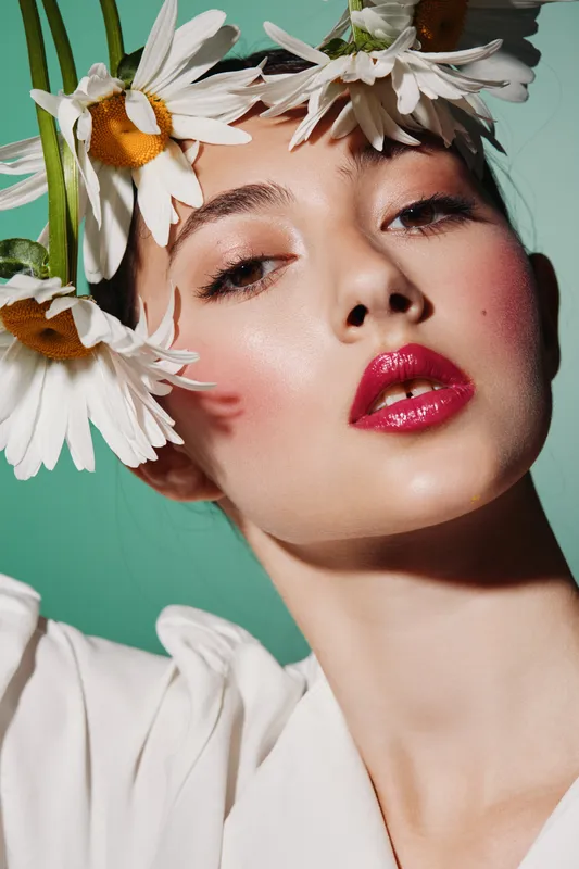 Close-up of a fashionable woman with glossy red lips and daisies in her hair against a pastel green background. Creative beauty concept with shining skin and intense gaze.
