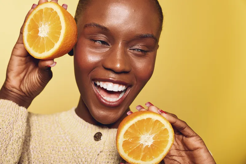 Happy African woman with dark skin holds two halves of fresh orange near face on yellow background. Concept of healthy lifestyle, vitamin C, skincare, natural beauty, and wellness.