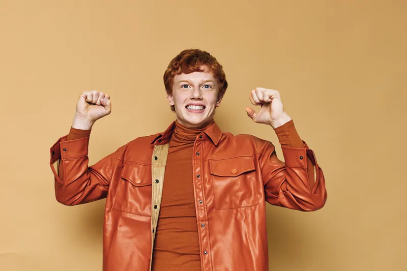 Young man with red hair and freckles smiles confidently with raised fists wearing orange shirt and jacket on bright beige background for success or celebration.