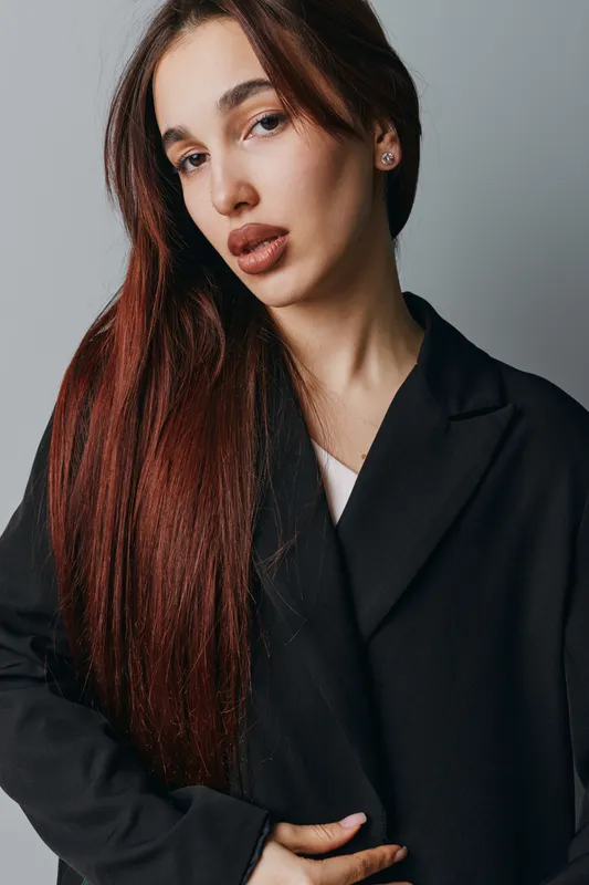 Young woman with long brown hair wearing black coat posing against gray background. Portrait of confident female model with natural makeup and serious expression in studio setting.