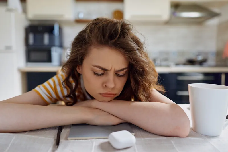 Young woman with wavy hair rests on a kitchen table, head on forearms, eyes closed. Home lifestyle scene conveying fatigue, stress, and quiet introspection under soft natural light.