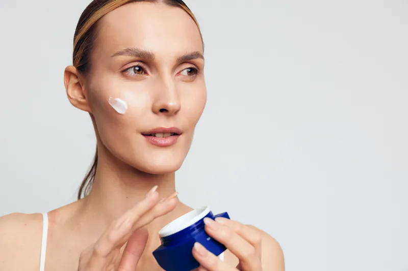Young woman with clear skin applies moisturizing cream on face holding blue jar isolated on white background skincare beauty routine concept natural makeup and healthy lifestyle
