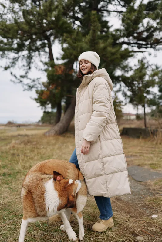Winter scene with a woman in a long beige coat and knit hat walking outdoors with a dog in a park, showcasing warmth, style and companionship