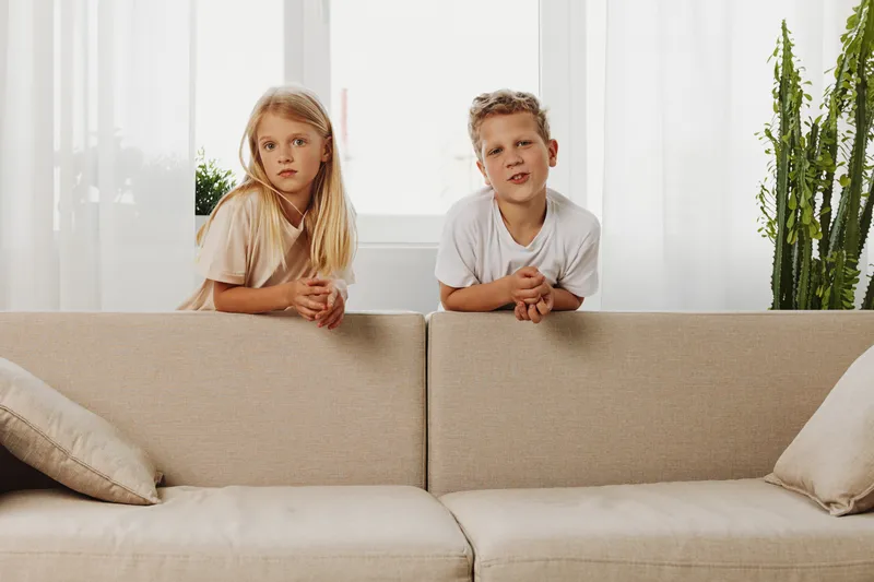 Two children lean on beige sofa in bright room with plants, looking curious and thoughtful. Concept of childhood, home, and family moments.