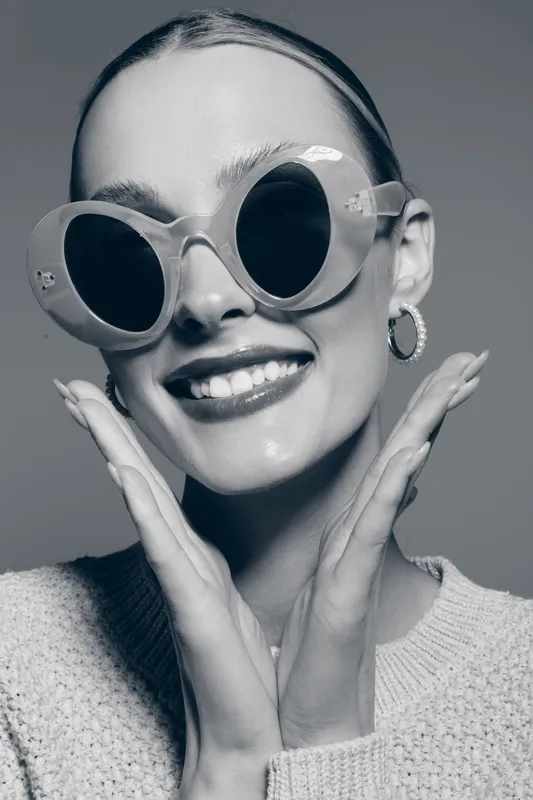 Smiling young Caucasian woman in round sunglasses poses with hands near face. Concept of fashion, beauty, and positive mood in studio portrait.