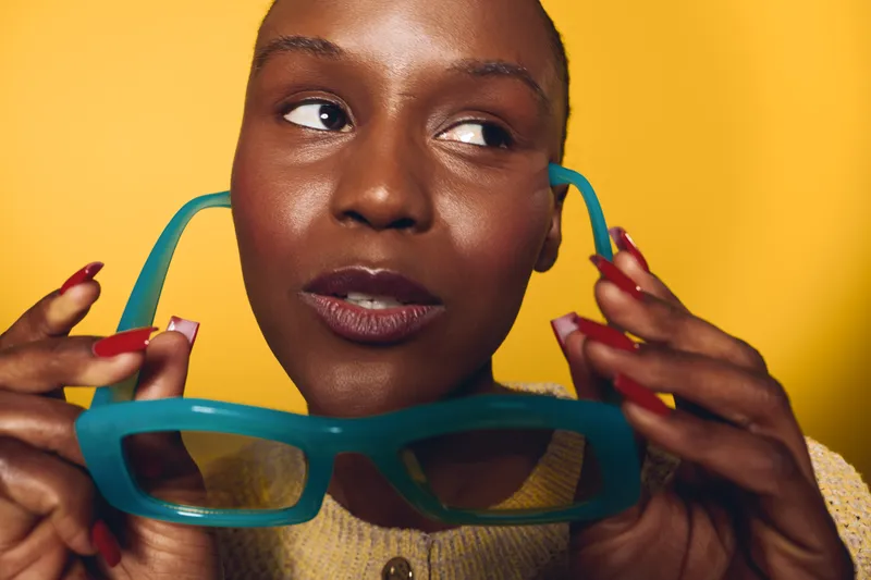 Close-up of confident dark skinned African woman holding blue sunglasses near face with thoughtful expression. Yellow background highlights natural beauty and modern style concept.