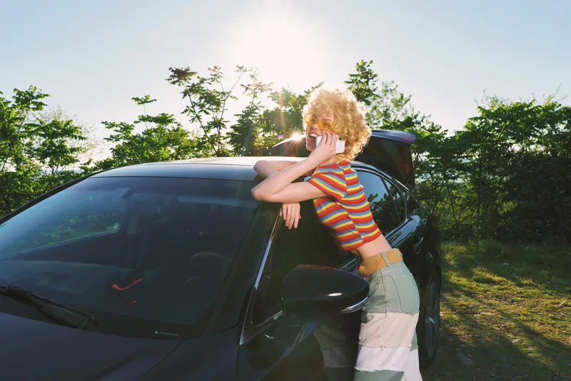Woman leaning on a car wearing a striped shirt and denim skirt, playful casual lifestyle shot in sunny outdoors, golden hour glow, relaxed mood, carefree summer drive moment.