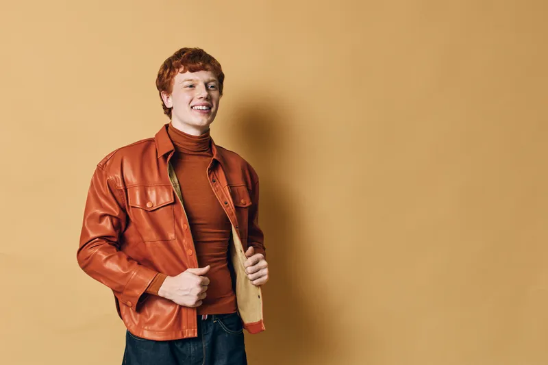 Young man with red hair wearing orange leather jacket and brown turtleneck posing smiling on beige background in casual style with hands in pockets.