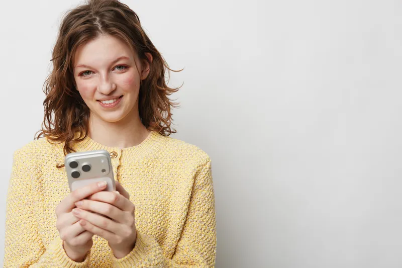 Smiling young woman in yellow sweater holds smartphone, looks at camera with friendly expression. Concept of communication, technology, social media, and casual lifestyle.
