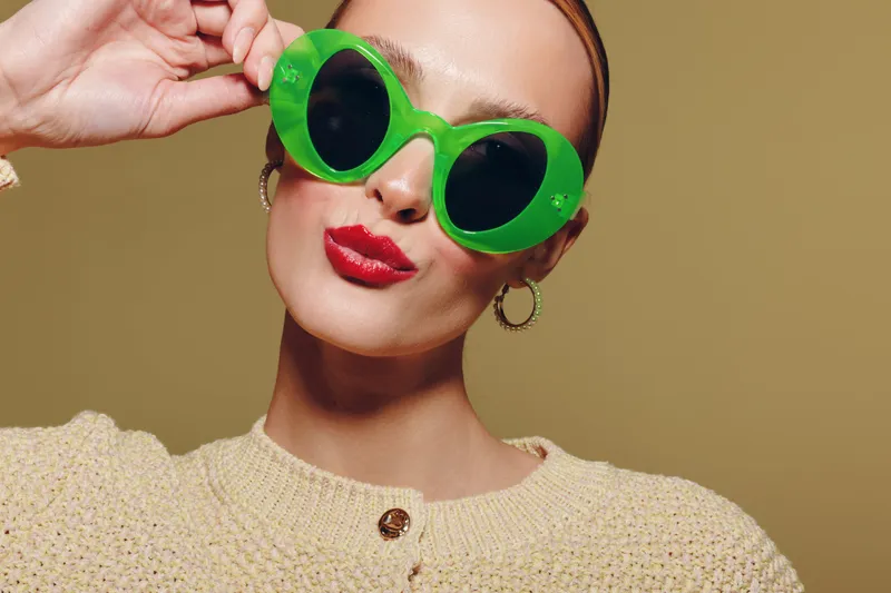 Stylish woman wearing large green sunglasses and red lipstick poses confidently against beige background. Fashion concept with bold accessories and modern makeup style.