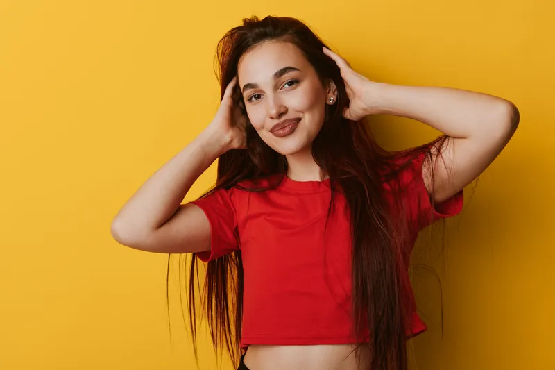 Young woman with long hair wearing red crop top posing with hands near head smiling confidently on yellow background. Female model with natural makeup and casual style in studio.