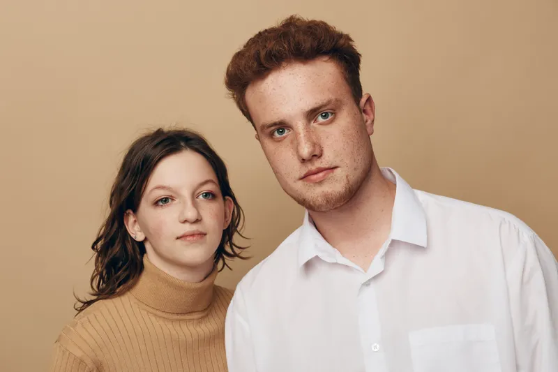 Young man and woman with serious expressions pose together against beige background. Casual clothing and natural look create a calm, neutral mood for portrait or lifestyle concept.