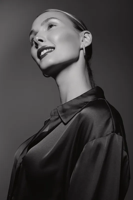Portrait of smiling caucasian woman with light skin and slicked hair wearing dark satin shirt on gray background.