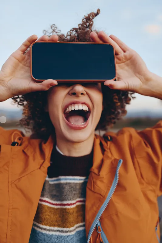 Woman lifestyle portrait outdoors, hands hold smartphone with blank screen, curly hair visible, she laughs open mouth, orange jacket, warm daylight, blurred background, happy mood today.