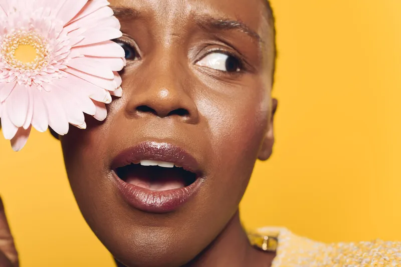 Close-up of a surprised dark skinned African woman holding a pink flower near her face. Yellow background highlights natural beauty, emotion, and floral concept with fresh skincare vibes.