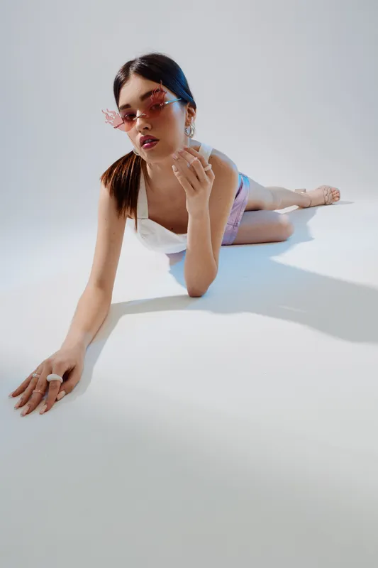 Young woman in stylish sunglasses and trendy outfit poses lying on the floor in a bright studio. Concept of woman beauty fashion showcasing modern style and attitude.