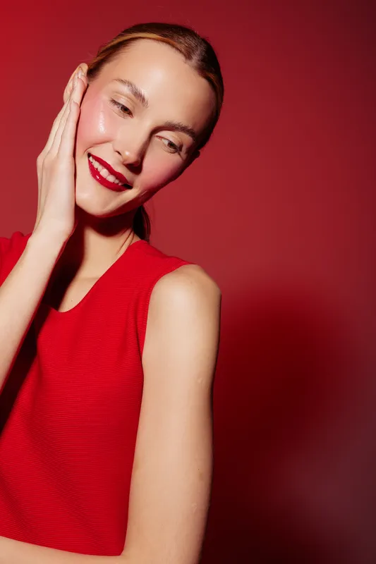 Smiling woman with wet skin and red lipstick wears sleeveless red top against red background. Concept of beauty, skincare, makeup, and fashion with vibrant color contrast and studio lighting.