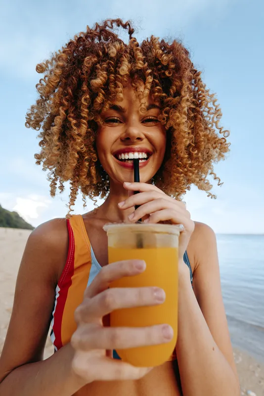 A joyful woman on the beach drinks bright orange juice, smiling at the camera. Outdoor summer scene, refreshment, vitality, and carefree coastal vibes.