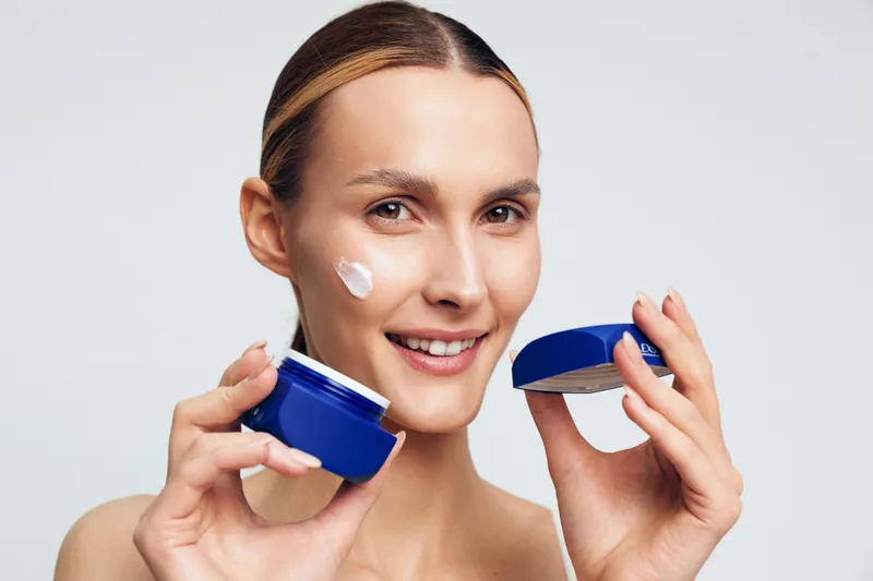 Young woman applying face cream with a smile, holding blue jar, clean skin, skincare routine, white background, natural beauty, closeup portrait, healthy lifestyle.
