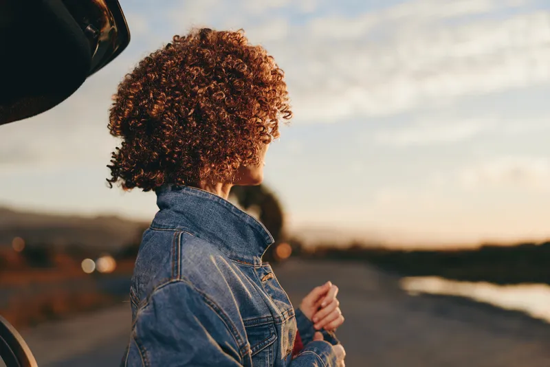 Woman lifestyle, curly hair, denim jacket, sunset, road, back view, outdoors, standing near car, soft golden light, travel mood, rear profile, highway, wind, walking, summer evening
