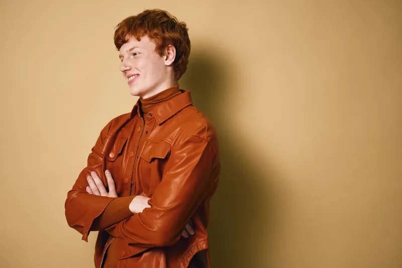 Young man with red hair wearing brown leather jacket standing with crossed arms looking sideways against beige background. Casual style with confident expression and modern outfit indoors.
