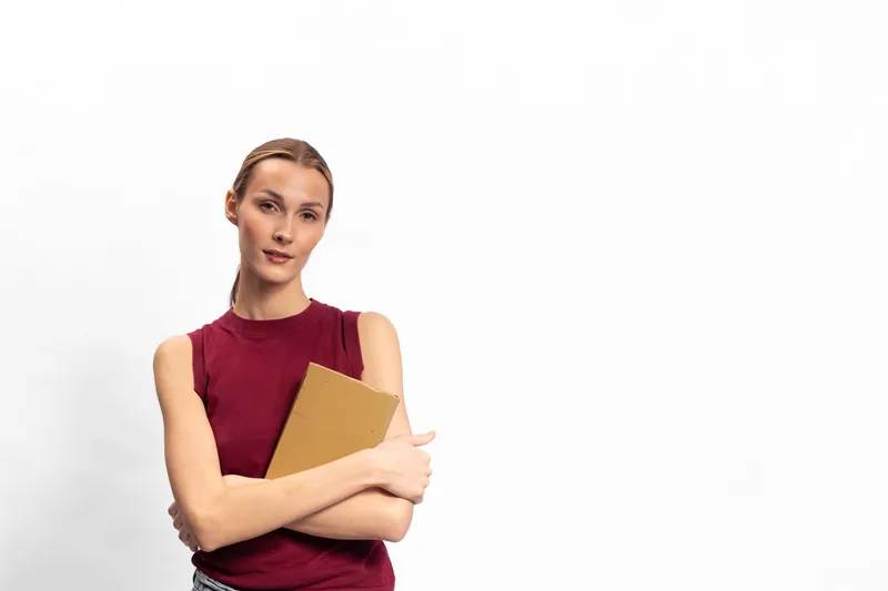 Confident young woman in red sleeveless top holds a notebook and looks at camera on white background. Concept for education, study, planning, and focus.
