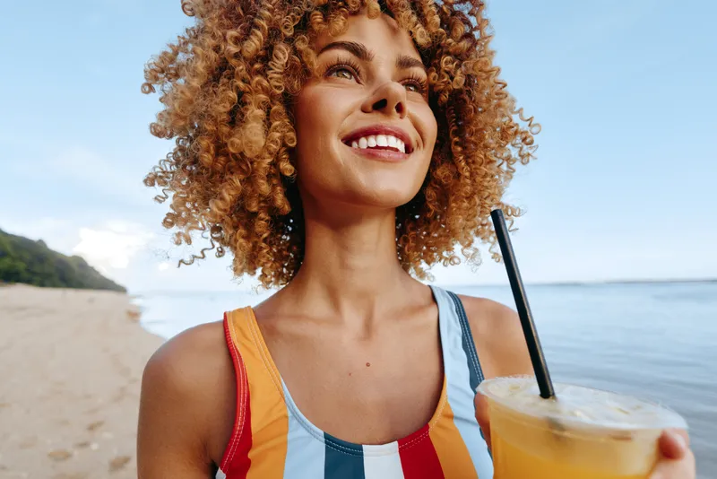 Young woman on a sunny beach laughs with a refreshing orange drink in hand, curly hair catching the breeze, striped top, calm ocean behind, bright summer moment