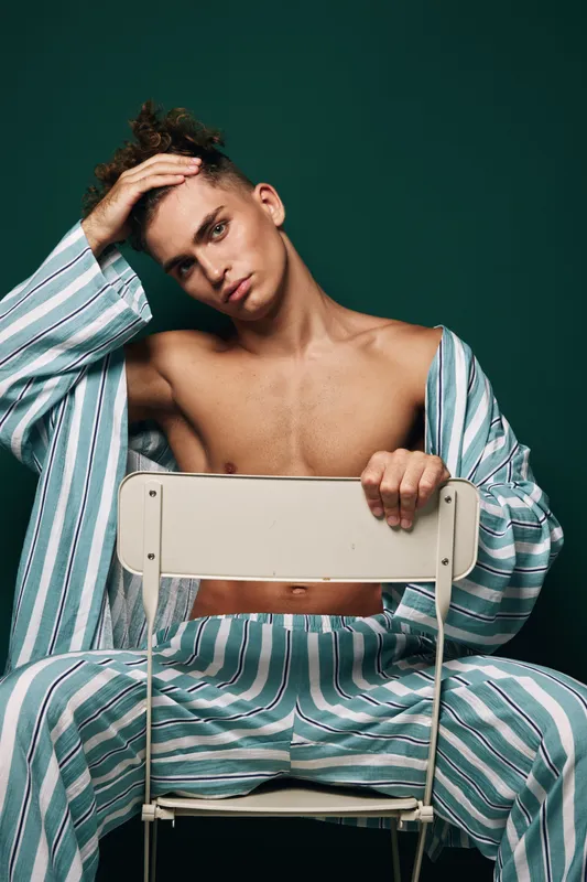 Young man in striped pajamas sitting on chair backwards with hand on head against green background. Casual relaxed pose with bare chest and thoughtful expression indoors.