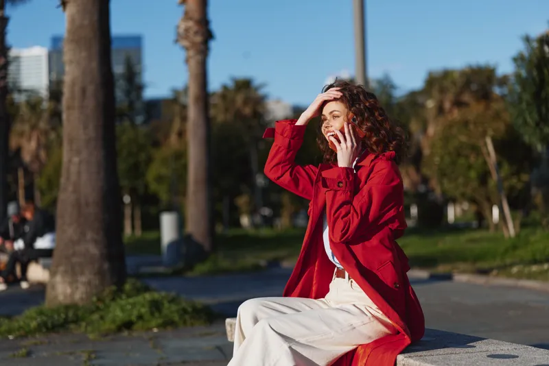 A young woman with curly hair sits on a bench wearing a red coat, shielding her eyes from the sun in a sunny outdoor park atmosphere.