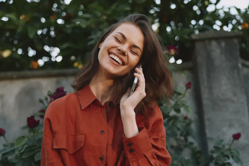 A cheerful woman in a red shirt chats on her phone outdoors in a garden, a candid lifestyle moment that conveys connection, happiness, and everyday communication