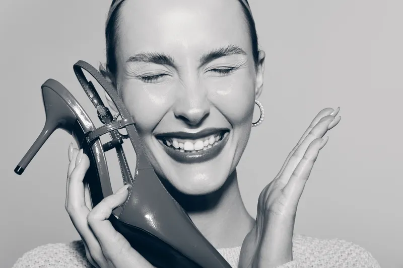 Happy white woman with pale skin laughs holding high heel shoe near face. Concept of fashion, joy, and modern style.