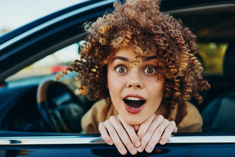 Young woman with curly hair leans from a car window, surprised and excited, capturing a candid street moment with vibrant colors and energetic vibe.