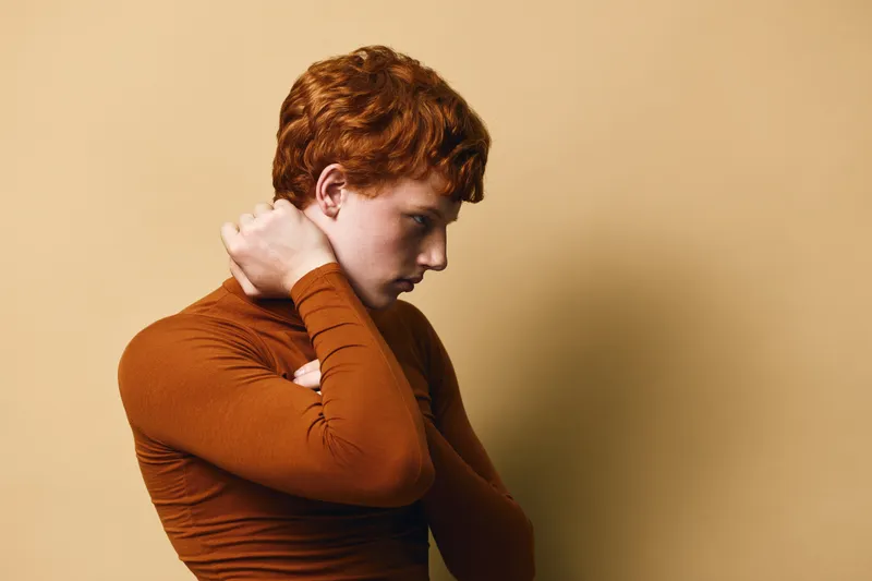 Young man with red hair and freckles wearing brown turtleneck sweater posing sideways with arms crossed on beige background. Portrait of thoughtful male in studio.