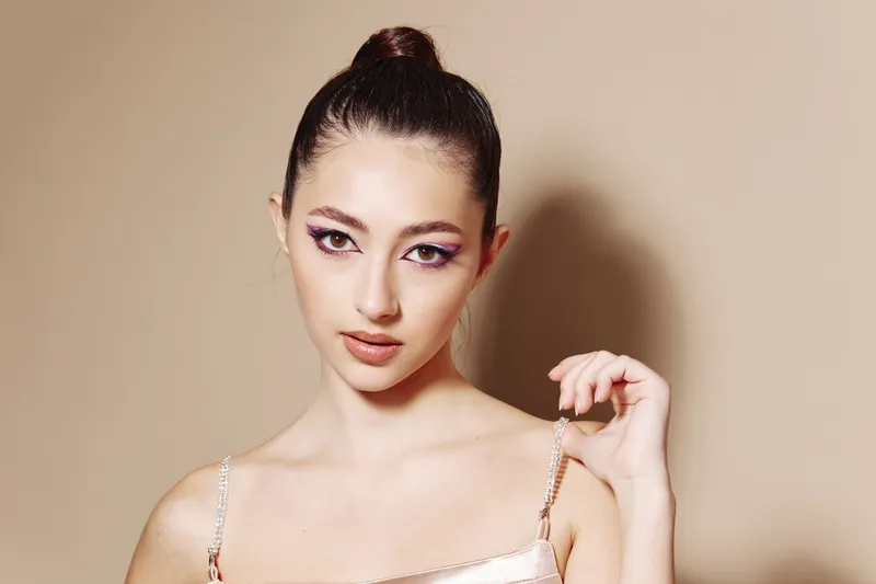Fashion woman with high bun hairstyle wearing shiny dress poses confidently on beige background. Creative beauty concept focused on glamorous makeup and stylish modern look.