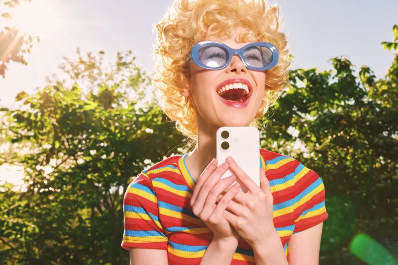 Joyful woman outdoors, laughing with eyes closed, sunlit moment, retro color tones, casual striped shirt, lush green background, carefree lifestyle