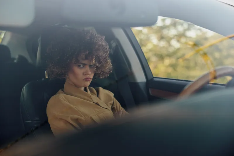 A woman sits in the driver's seat of a car, looking serious with a distant gaze, while warm daylight fills the quiet interior, capturing a moment of reflection during a city drive.