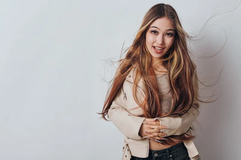 Young woman with long hair wearing casual jacket stands smiling with crossed arms against white background. Female model with natural look posing for portrait photography.