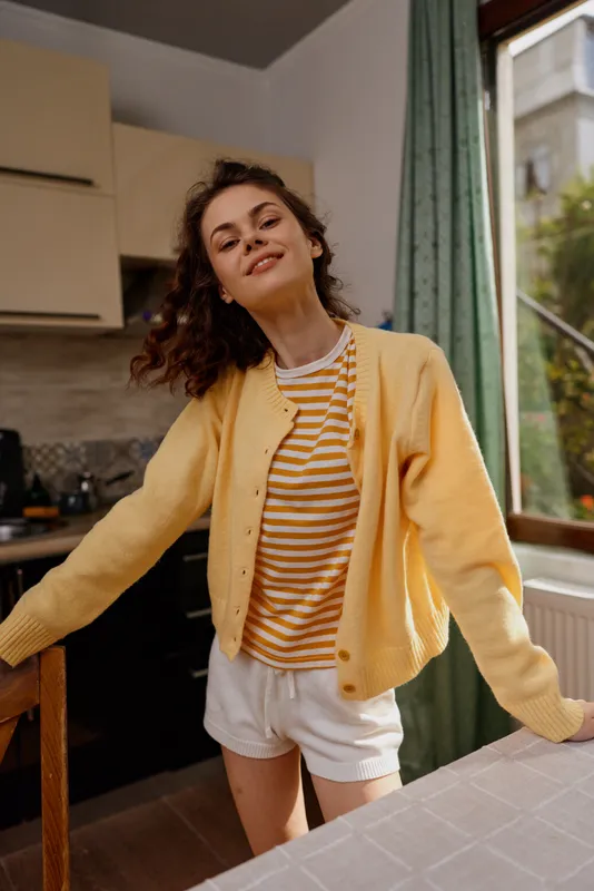 Young woman in a yellow cardigan and striped tee smiles and stretches in a sunlit home kitchen, creating a bright, carefree lifestyle moment.