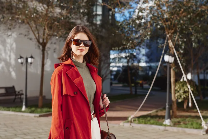 Stylish woman in a red coat walks outdoors in a sunlit urban plaza, wearing sunglasses and a green turtleneck, exuding confident chic and relaxed poise today