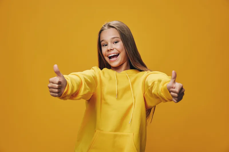 Girl in yellow hoodie smiling and showing thumbs up with both hands on yellow background. Happy teenager with long hair expressing positivity and approval in studio portrait.