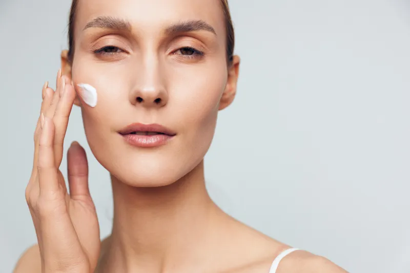 A closeup of a woman applying cream to her cheek in a bright studio, showcasing skincare routine and self care, with calm expression and clear skin.
