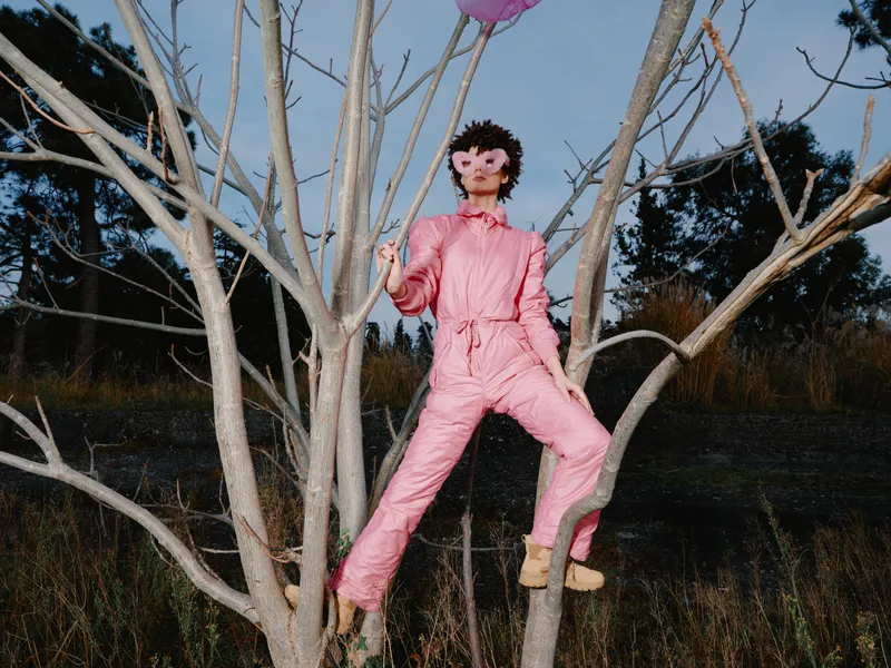 Person in a pink jumpsuit climbs a bare tree at night, creating a bold surreal scene with dramatic lighting and open landscape, highlighting climbing, creativity, and outdoor exploration