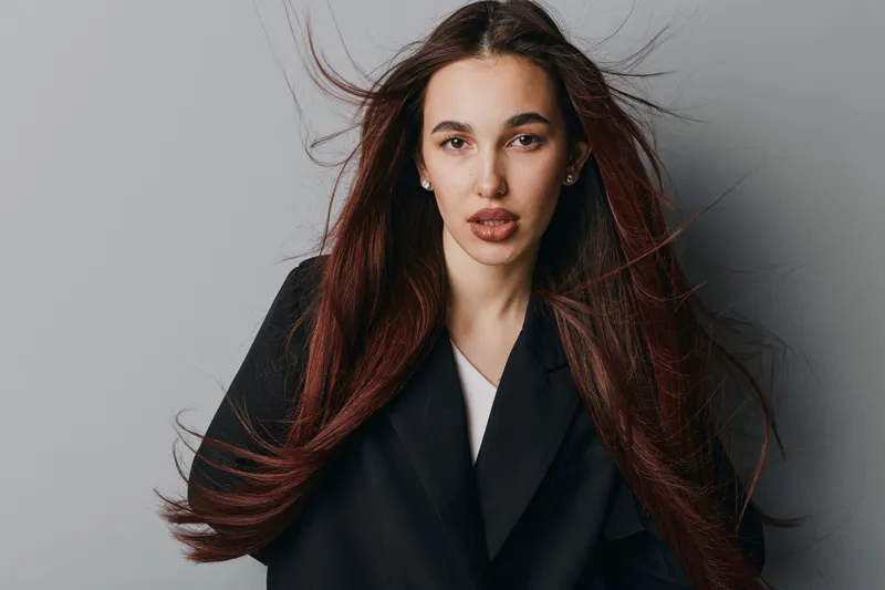Portrait of young woman with long flowing hair wearing black coat standing against gray background. Female model with natural makeup and serious expression looking at camera indoors.