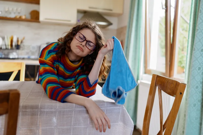 Relaxed woman with colorful striped sweater rests her head on her arm at a kitchen table, towel in hand. Casual home setting conveys comfort, wellness, and everyday life.