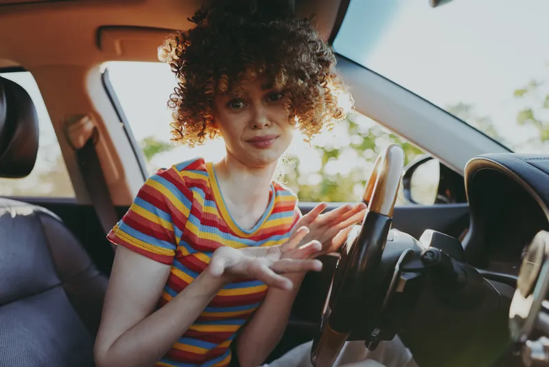 Curly haired woman in a striped tshirt sits in a car seat, gesturing with her hands. Bright natural light highlights casual travel and everyday moments.
