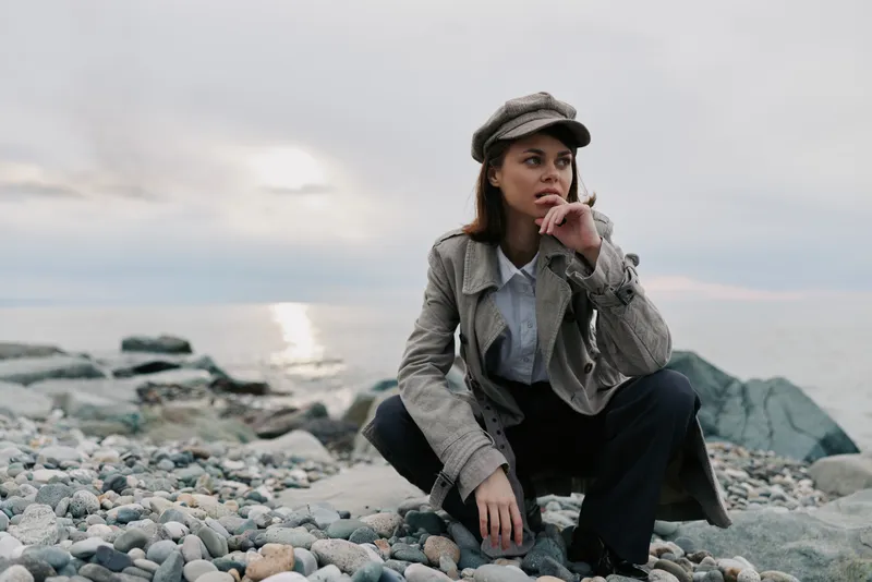 Young woman in a coat and hat squats on a rocky beach near the sea. Overcast sky and calm water create a moody atmosphere for outdoor fashion concept.