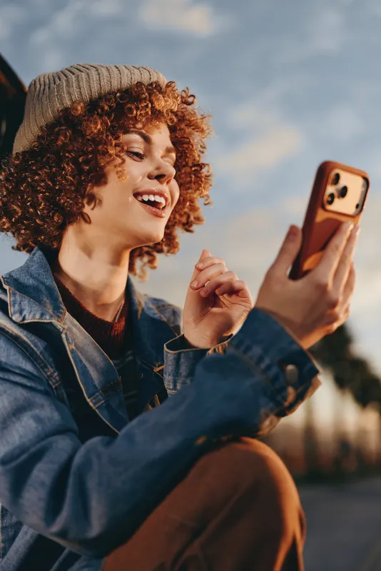 Woman outdoors, smiling with curly hair, holding a smartphone, denim jacket lifestyle, sunny sky, joyful casual moment, soft light, relaxed hand gesture, blurred urban background bright warm glow