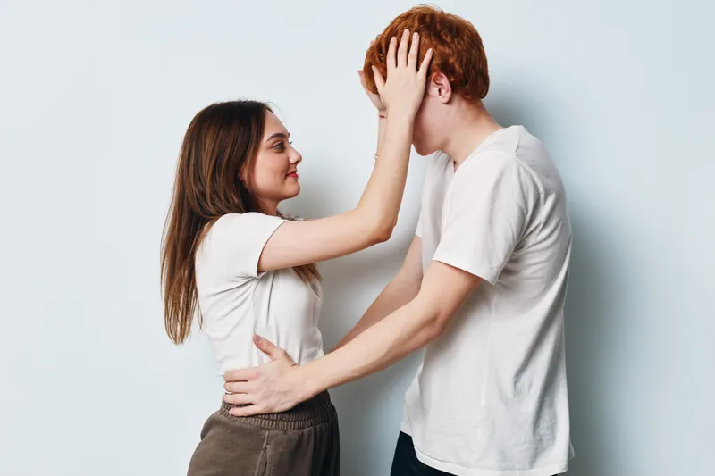 Young woman and man standing close, smiling and touching faces in affectionate pose against white background. Casual clothing with emotional connection and intimate moment of care.