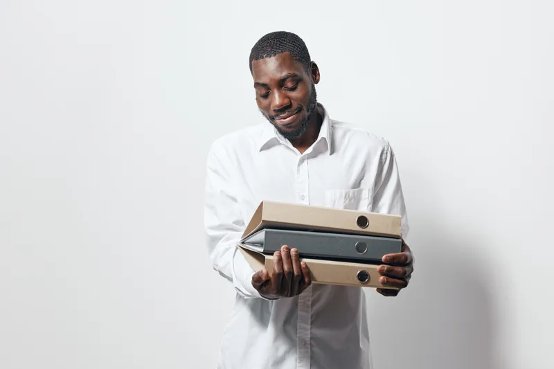 African American man with dark skin wearing white shirt holding office folders and binders, standing against white background with slight smile and professional posture.