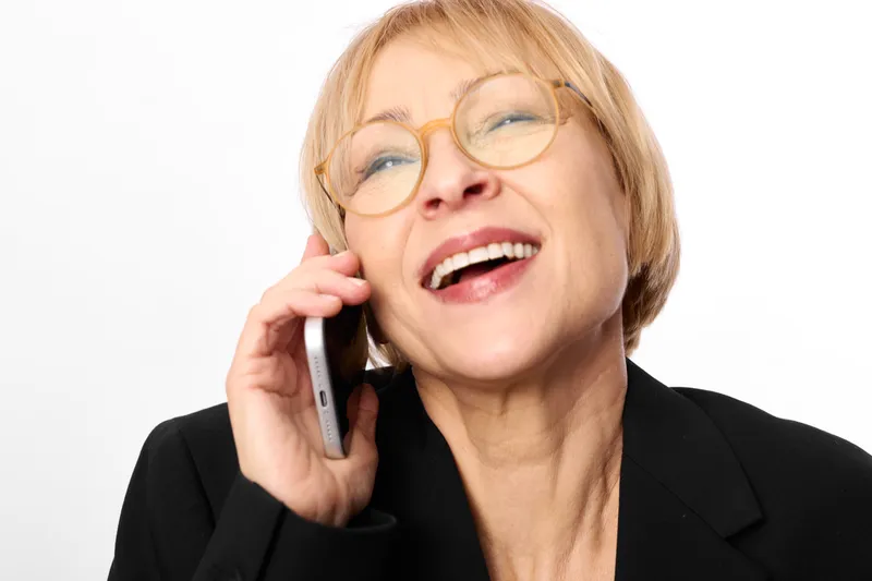 Elderly woman with glasses smiling while holding a phone to her ear in a studio portrait on a white background Business communication call